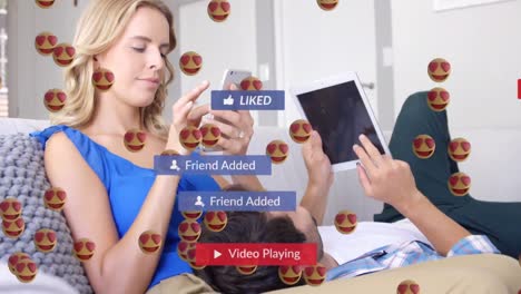 animación de ojos de corazón cara emojis flotando sobre pareja caucásica usando dispositivos electrónicos en el hogar.