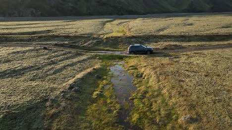 An off-road vehicle crosses a small stream in a remote valley in northern Iceland, surrounded by rugged terrain and untouched wilderness under clear skies.