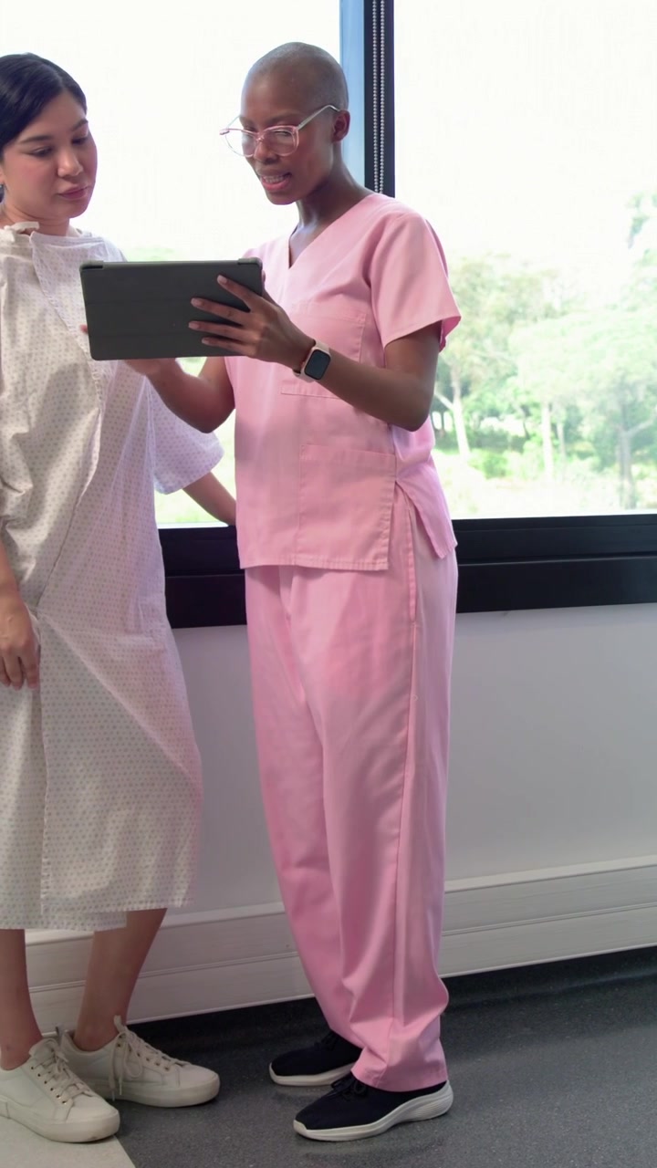 Video vertikal: Teknisi bergerak mengenakan seragam medis merah muda dari unit mammogram, mempersiapkan pasien bergaun dengan tablet.