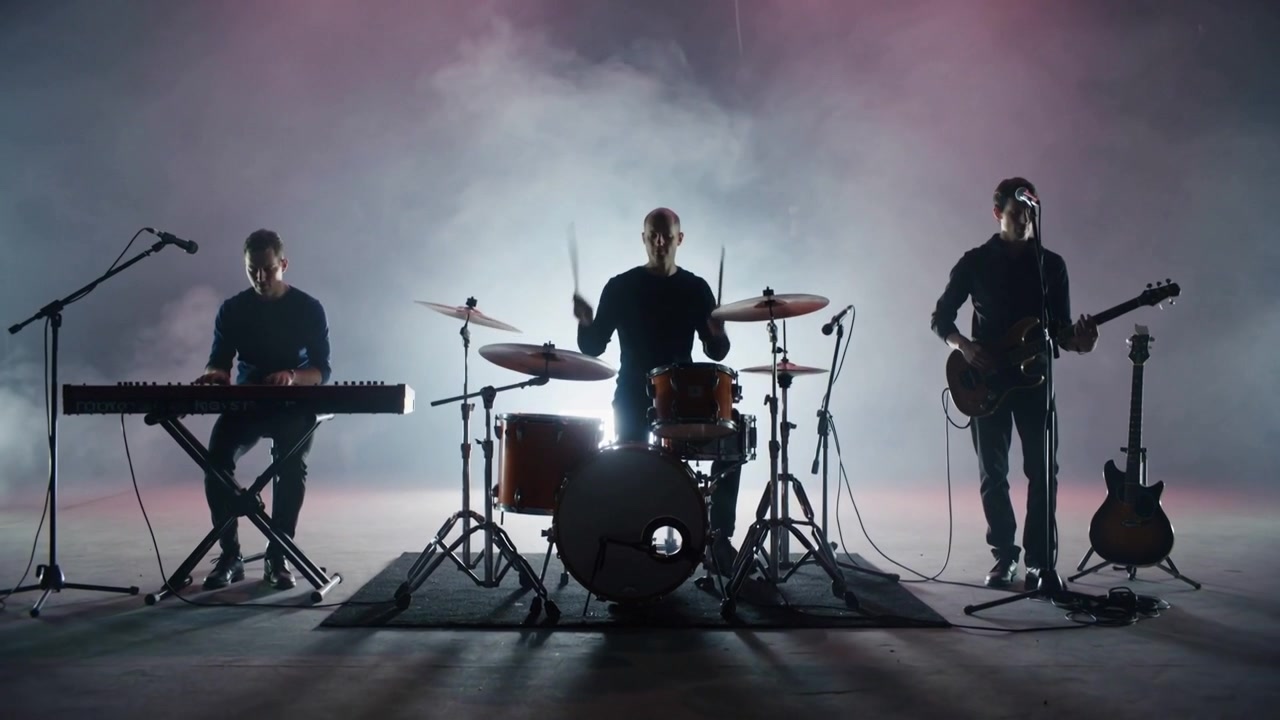Trio musicale che si esibisce per una registrazione su un palco di studio fumoso e retroilluminato, con abiti scuri, batteria, tastiere e chitarra.