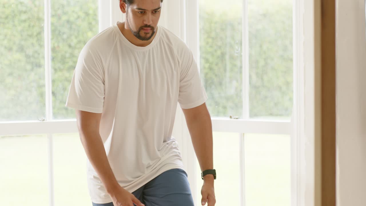 Uomo in equilibrio su una gamba, che tira su il ginocchio, riscaldandosi in maglietta a casa con i manubri