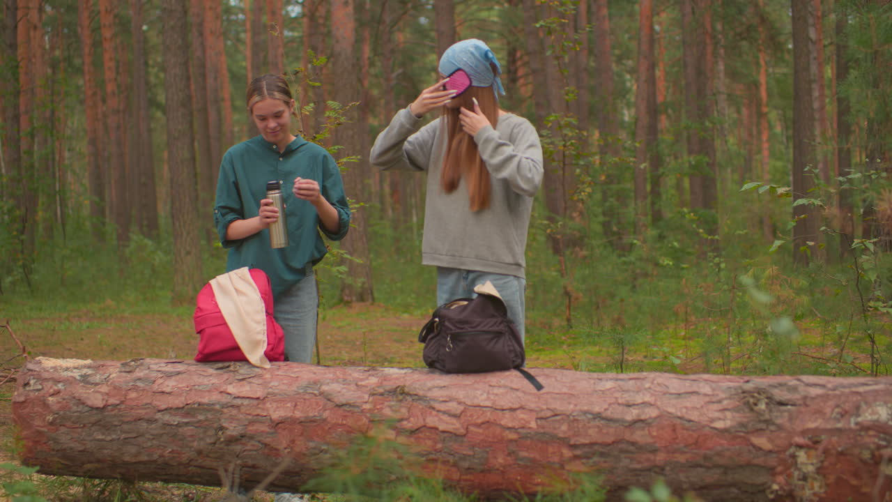 los excursionistas hacen una pausa en el bosque sereno, uno presiona la cubierta interna del termo para abrirlo, mientras que otro suavemente cepilla su cabello, comparten una sonrisa cálida, las mochilas descansan en el árbol caído