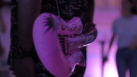 a man wearing a pink balloon guitar at a party with people dancing in the background - close up