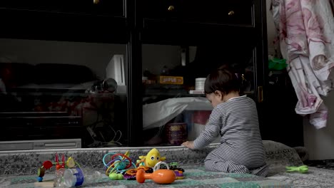 8-Month-Old-Baby-Wearing-Babygrow-Trying-To-Open-TV-Cabinet-Glass-Door-With-Toys-On-The-Floor-In-Living-Room