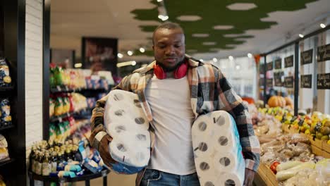 retrato de un hombre feliz y alegre con color de piel negra en una camisa a cuadros y auriculares inalámbricos rojos que lleva grandes rollos de toallas blancas desechables mientras camina y baila cerca de vitrinas en un supermercado moderno