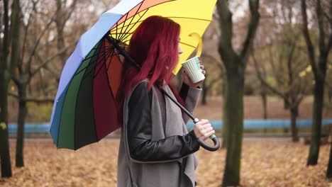 side view: young woman with red hair walking in autumn park and drinking coffee from a paper cup while holding colorful umbrella. girl in warm coat enjoying cool fall weather with a cup of hot drink