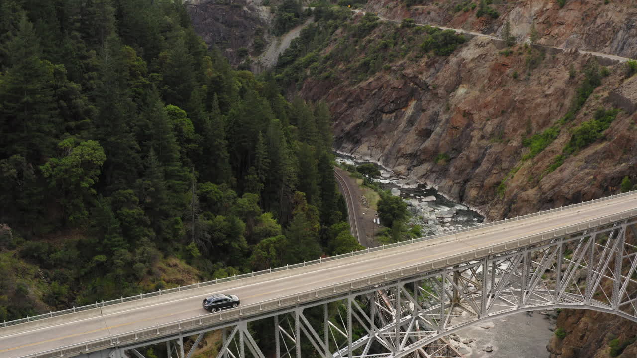 Drone pull back view going over an extremely tall metal bridge spanning another smaller bridge and a river. Cars pass on bridge.