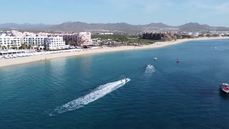 Aerial-view-in-front-of-the-coast-of-medano-beach-in-cabo-san-lucas,-mexico-with-view-of-the-floating-boats-in-the-blue-sea,-hotel-facilities-for-tourists-and-beautiful-mountain-landscape