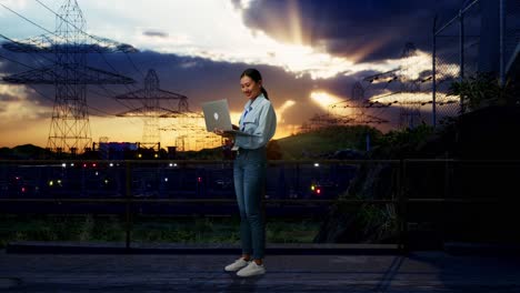 vista lateral completa del cuerpo de una trabajadora profesional asiática de pie con su portátil cerca de la torre de alto voltaje, instalación industrial, escribiendo en el teclado de su portátil con meditación