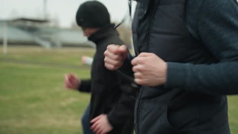 close-up of three people jogging on a track near a grassy field, with a clear view of the adult wearing a whistle in the foreground and a blurred view of the other young boys in the background