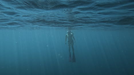 female freediver going up in epic wide oceanic shot, sunny light rays through water surface