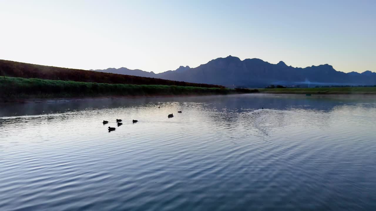 A cinematic drone soars smoothly across the glassy lake surface, its wake subtly distorting the perfect reflection of mist-shrouded mountains in golden dawn light