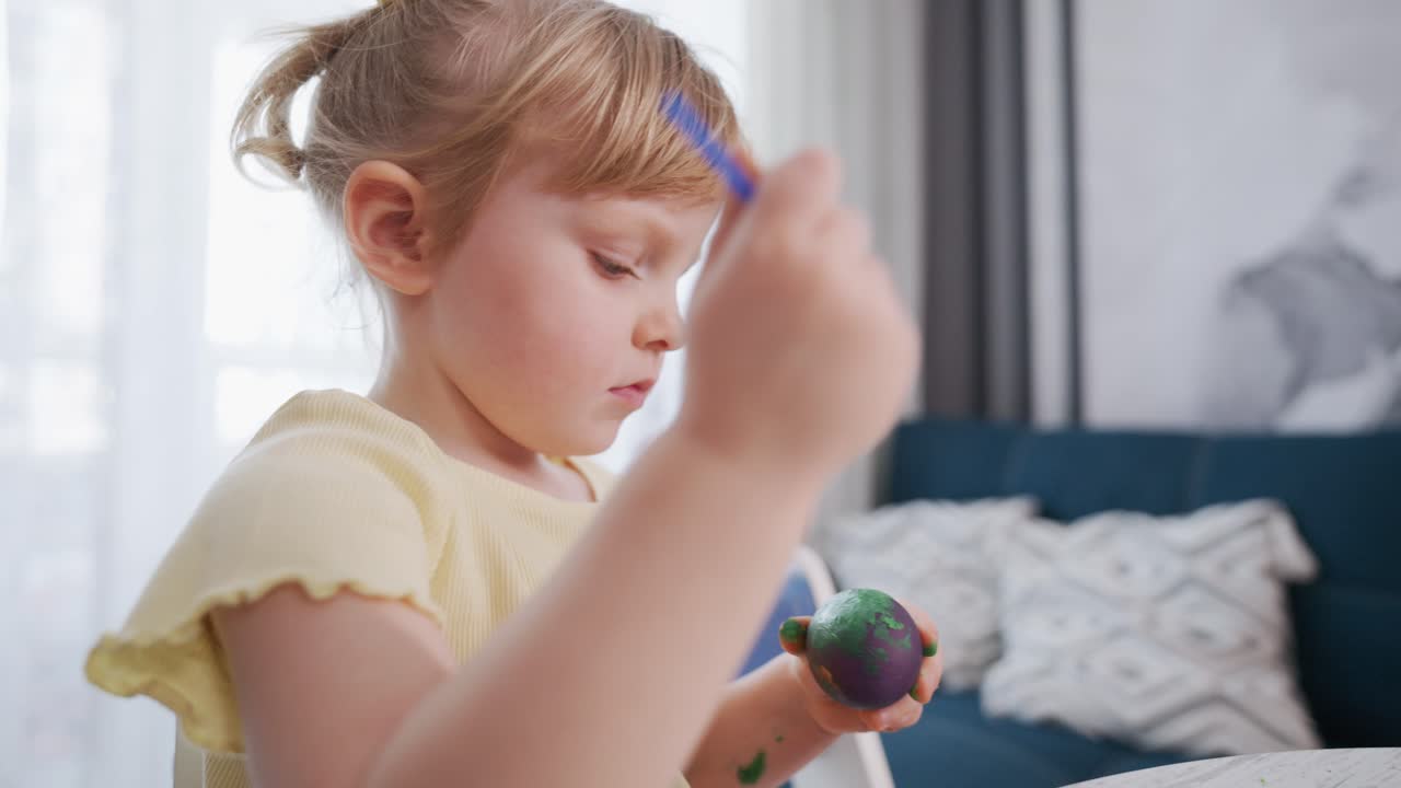 Young girl paints colorful eggs with focused attention. She wears a yellow shirt and has her hair tied in a ponytail. The scene is bright, indoors, near a window and sofa