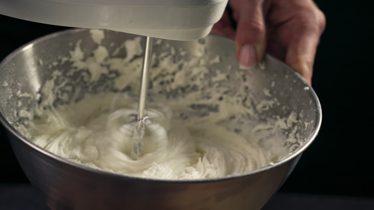 Hand holds bowl and mixer at angle to combine all ingredients into a whipped frosting, slow motion