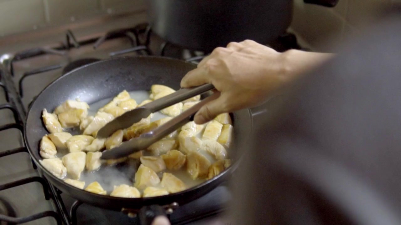 Close up shot of a person using metal tongs to stir and cook diced chicken pieces in a simmering lemon sauce on a gas stove. Authentic home cooking or restaurant kitchen scene.