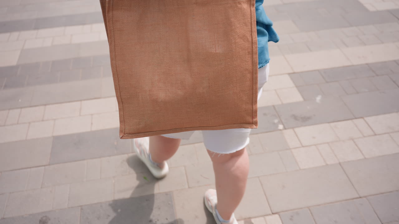 Close up rear view of woman walking on paved walkway in bright daylight carrying brown tote bag, dressed in casual blue shirt and white shorts, showing confident stride