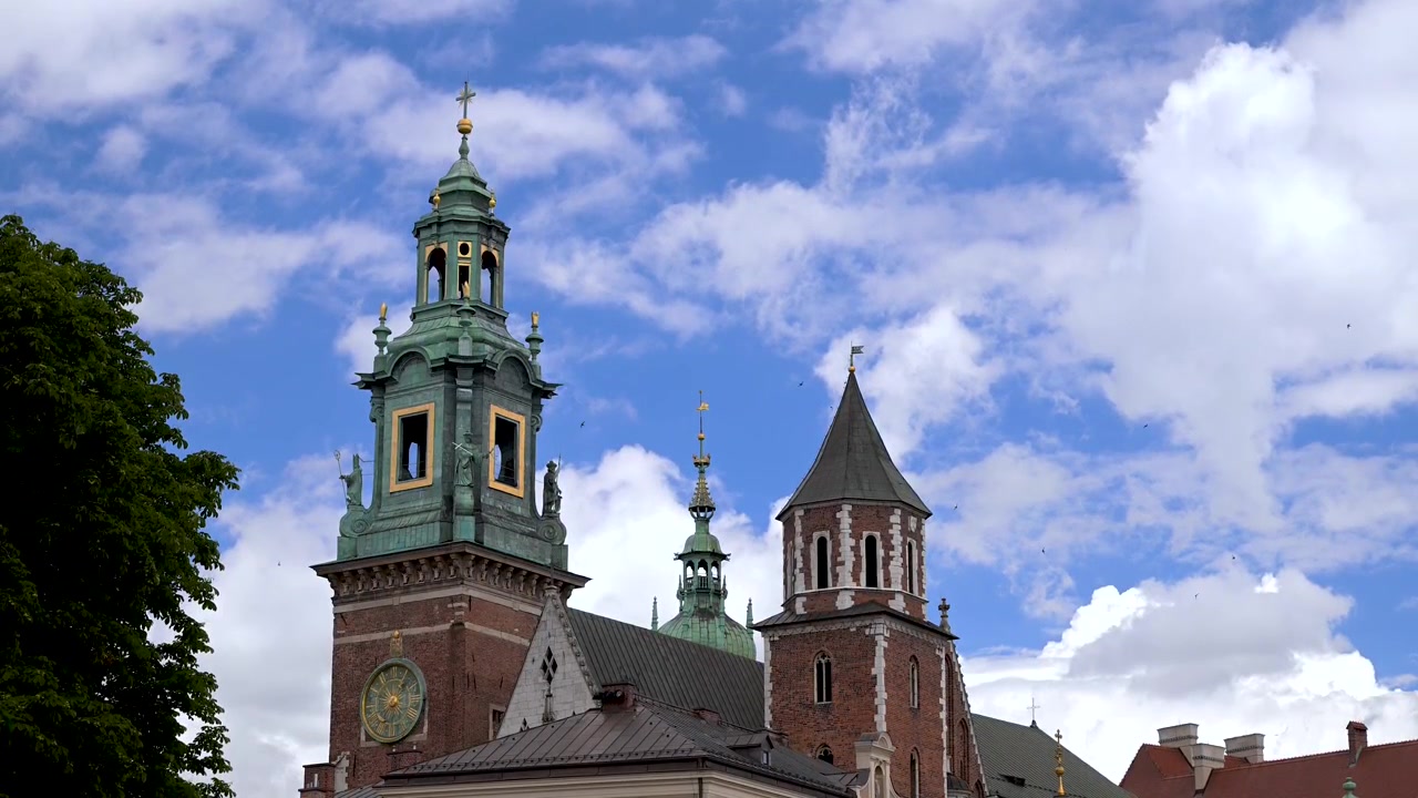 I campanili in mattoni della Cattedrale di Wawel e la guglia in rame verde si ergono sopra i tetti sotto un cielo estivo luminoso con nuvole alla deriva