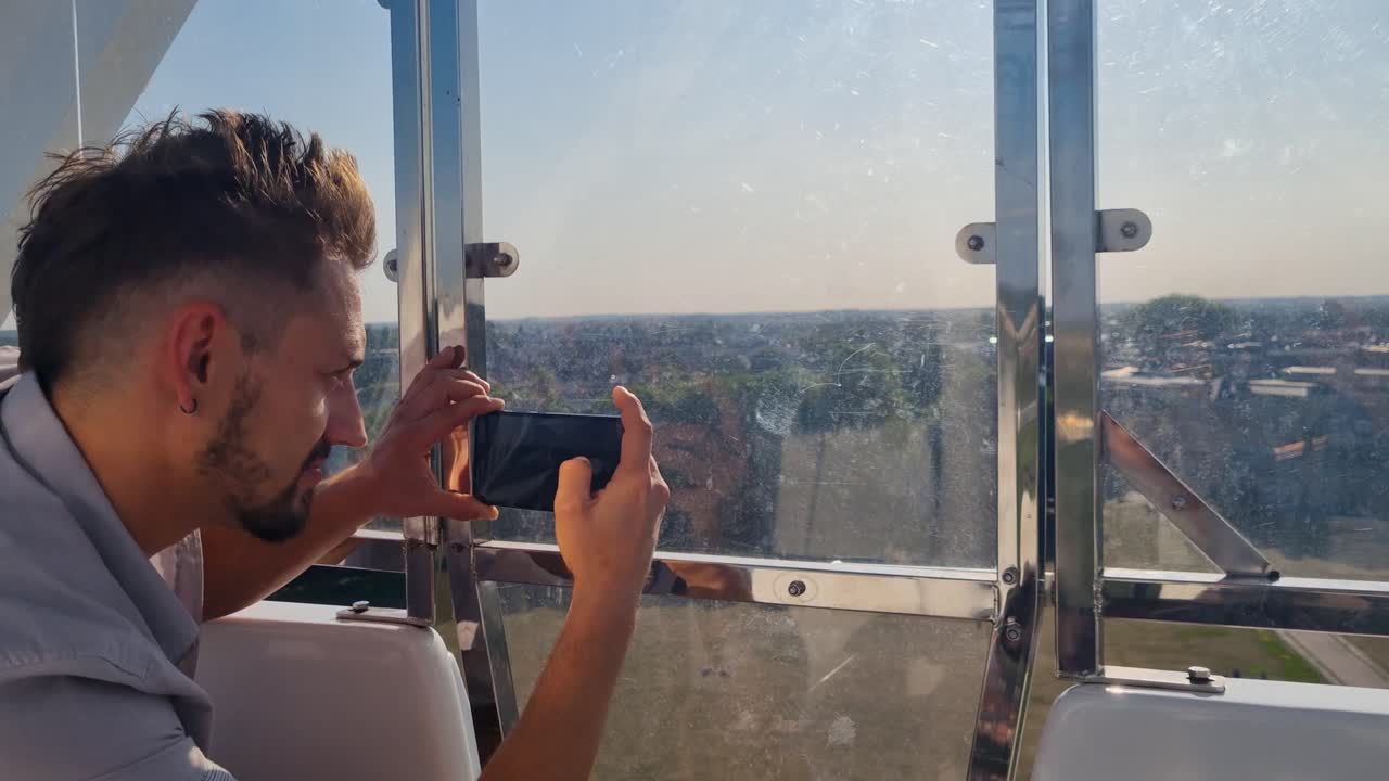 A man photographs Cambridge, UK, from a Ferris wheel cabin on a sunny day, capturing city rooftops and streets from a high, scenic vantage point