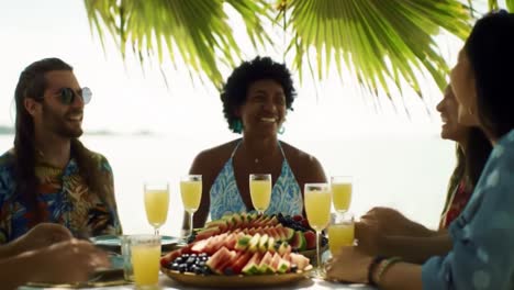 a joyful gathering by the sea: friends sharing laughter, fresh fruits, and refreshing drinks under a palm canopy in an idyllic setting