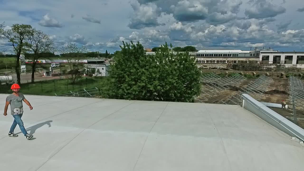 Technician in orange helmet on the roof. Worker goes on a flat roof of industrial building on the background of a construction of a new solar farm. Aerial view.