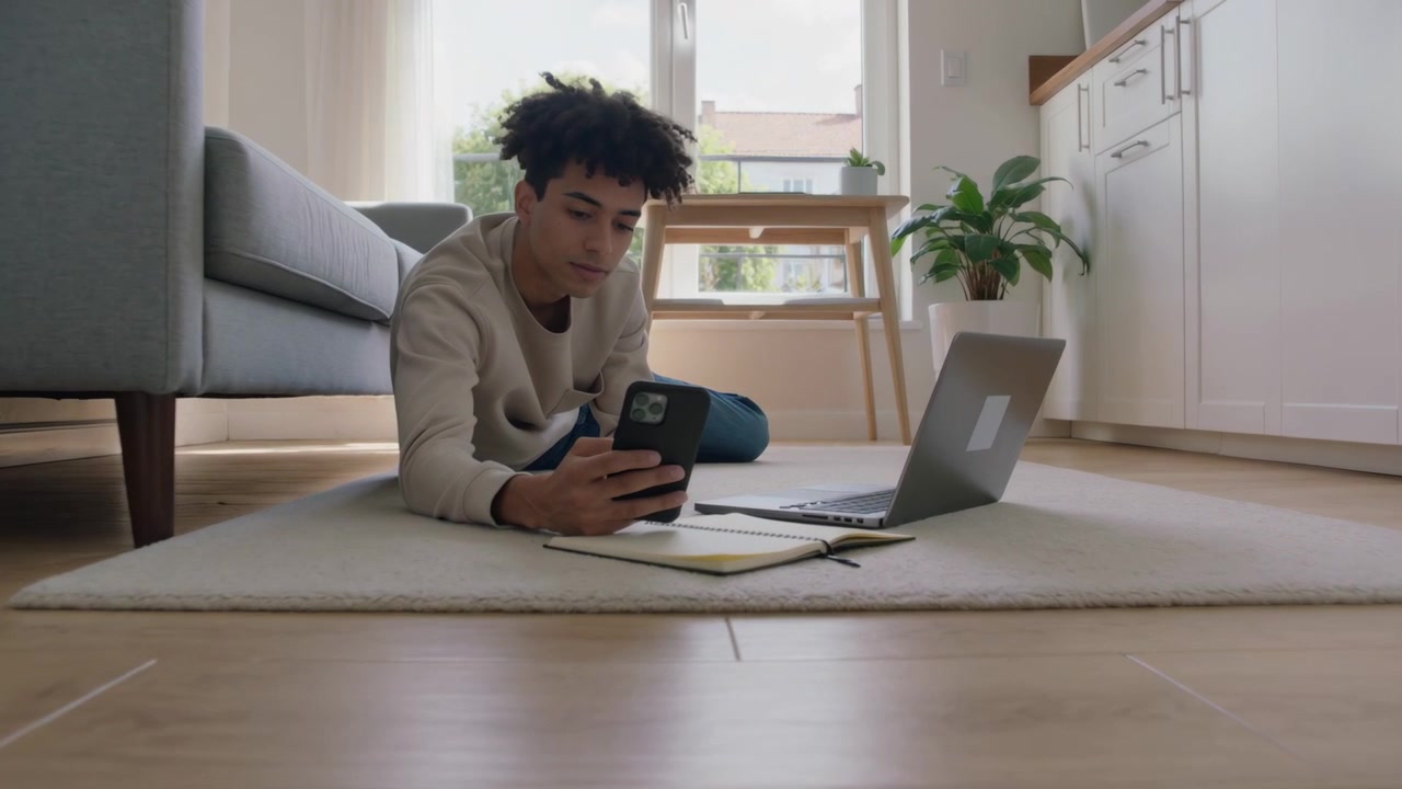 Propping on elbows, teen scrolling smartphone, glancing at laptop while studying on rug, notebook