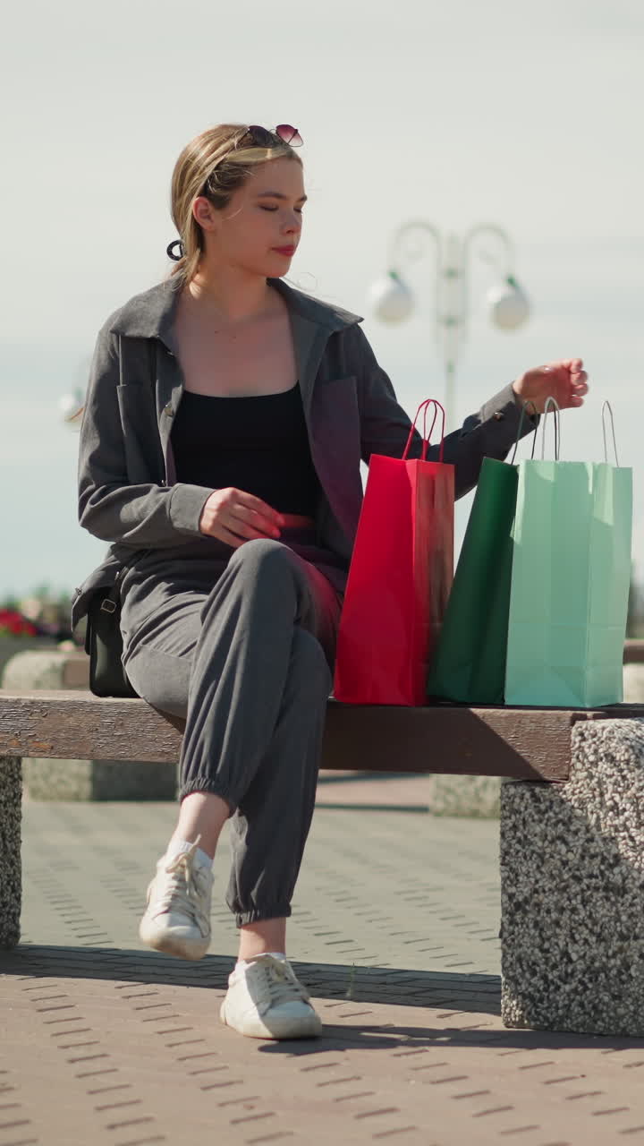 dama de ropa gris deja caer su bolsa de compras en un banco al aire libre, ajusta una bolsa de compras roja mientras sostiene un bolso de mano negro se sienta en el banco, mira hacia la derecha con una expresión enfocada