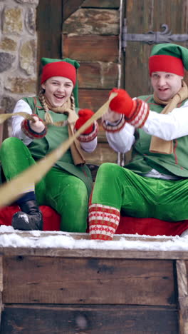 two elves in green costumes sitting in wooden sleigh outdoors. happy elves sit together in old fairy sleigh on the house background in winter. christmas time. vertical video