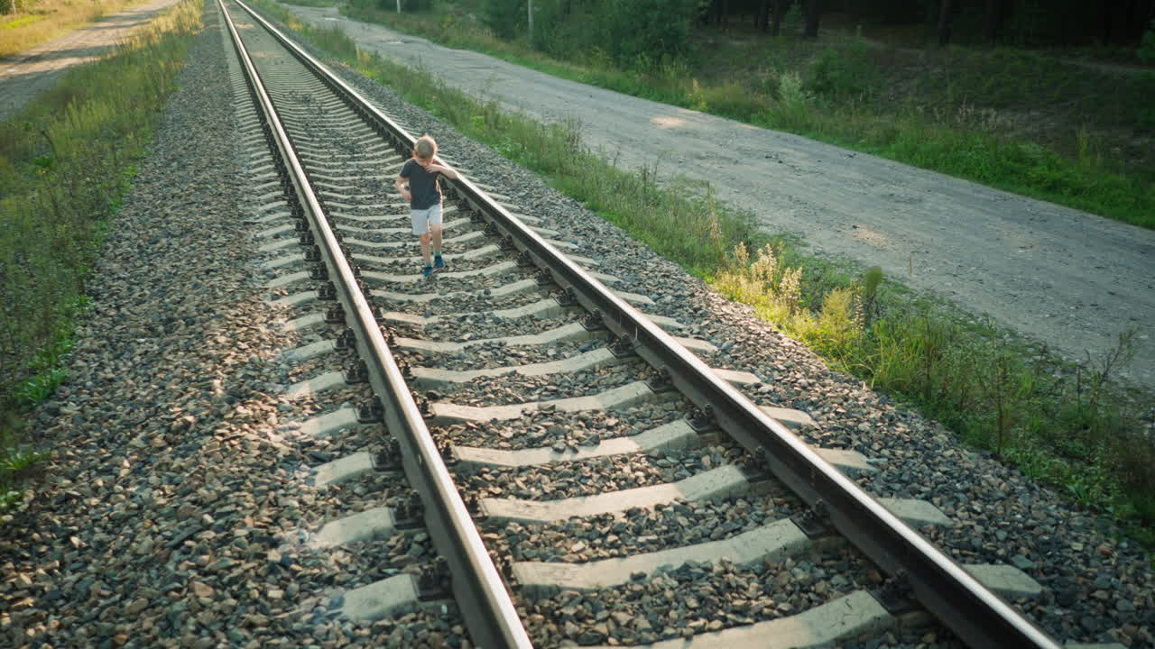 little boy in gray t-shirt and light shorts walking alone on rail track in rural area, wiping sweat from forehead on warm sunny day with gravel path and green vegetation on both sides