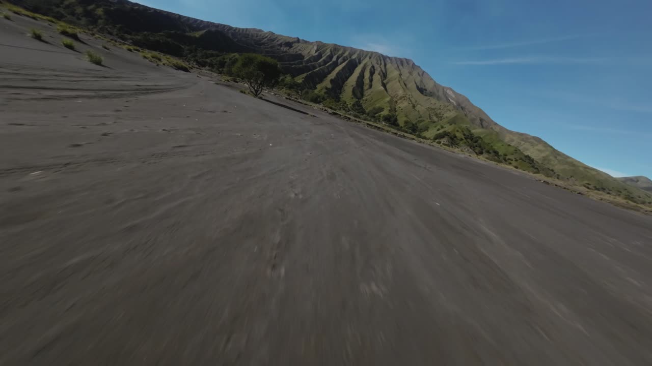 Riprese dinamiche con drone FPV che sorvolano a bassa quota il Mare di Sabbia del Bromo verso il vulcano Monte Bromo a Giava Orientale, Indonesia, mostrando un drammatico paesaggio desertico vulcanico.