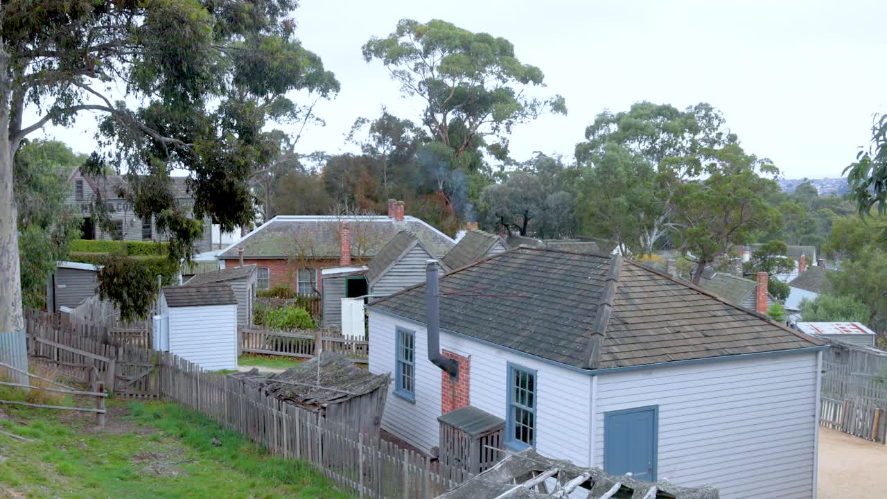 A serene view of a wooden house with smoke rising from the chimney, surrounded by greenery in Melbourne's Sovereign Hill
