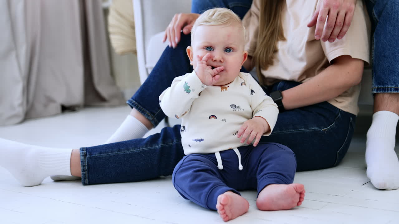 Adorable blond baby boy with grey eyes sits on the floor and chews his fingers. Unrecognized parents sit at backdrop near their child.