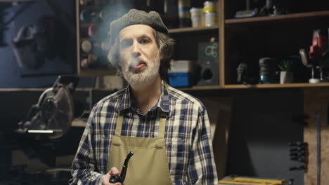 portrait of old caucasian handicraftsman in hat and apron smoking tobacco pipe while standing in carpentry workshop