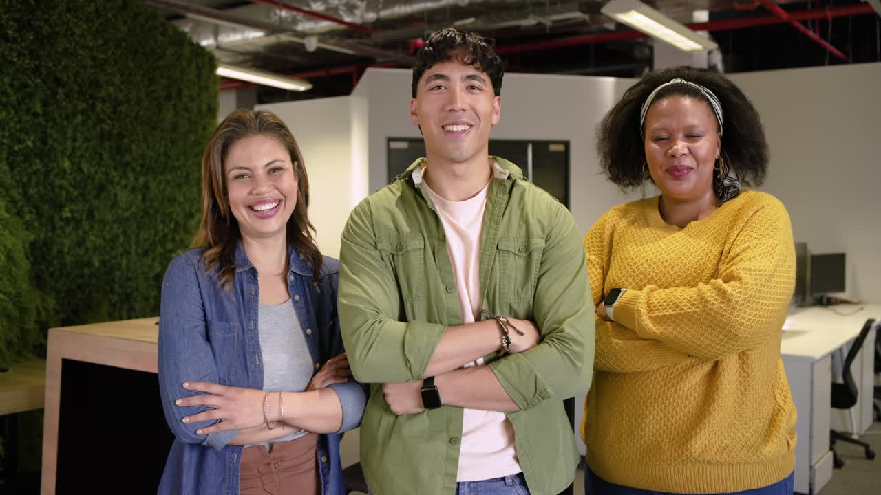 Tres compañeros de trabajo frente a la cámara, sonriendo, riendo, cruzando los brazos y proyectando confianza en la oficina.