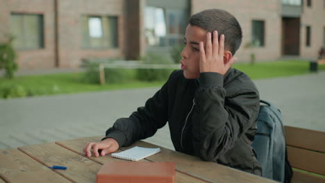 frustrated boy slams pen on notebook during study session outdoors, resting head on hand in visible irritation, sitting at wooden bench with book and backpack beside him in urban environment