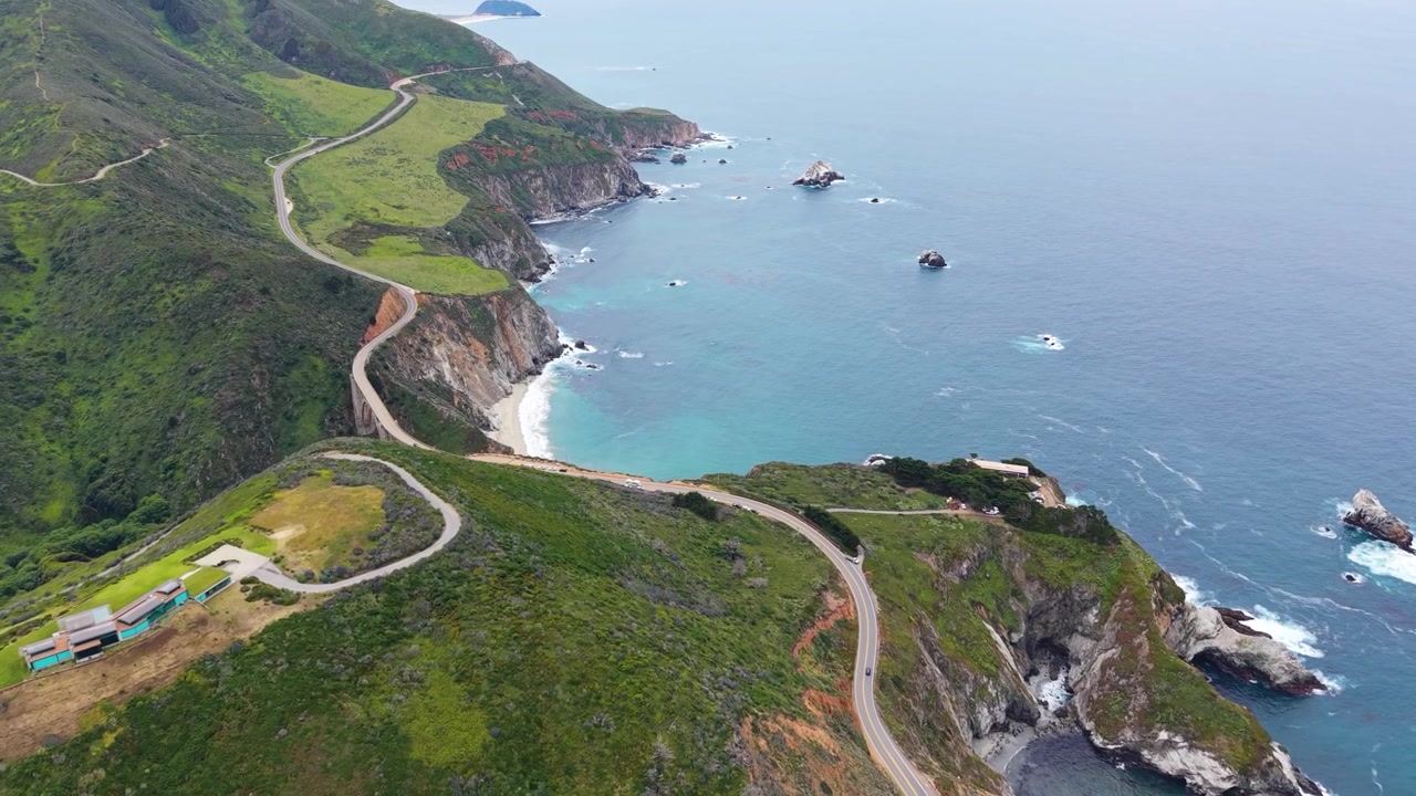 Aerial drone view of Highway 1 winding along steep green cliffs above turquoise water and offshore sea stacks in Big Sur.
