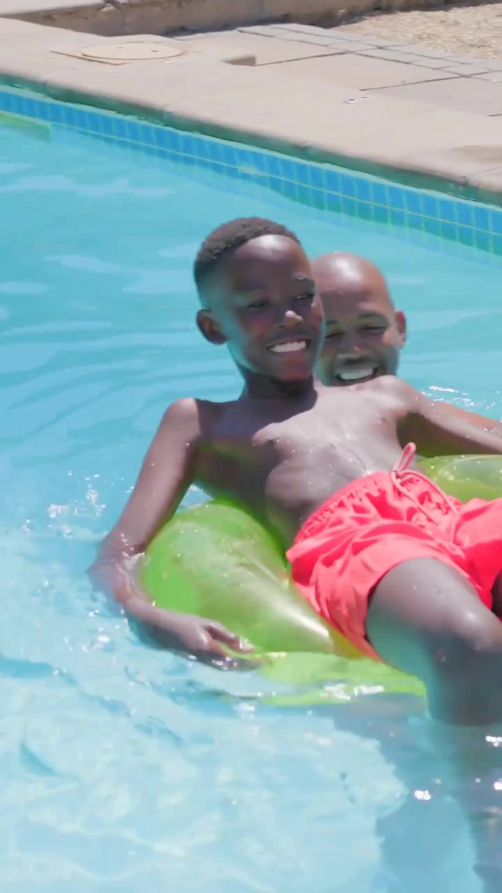 Vertical video: Helping dad steadying son in red trunks boarding green tube at pool, learning swim