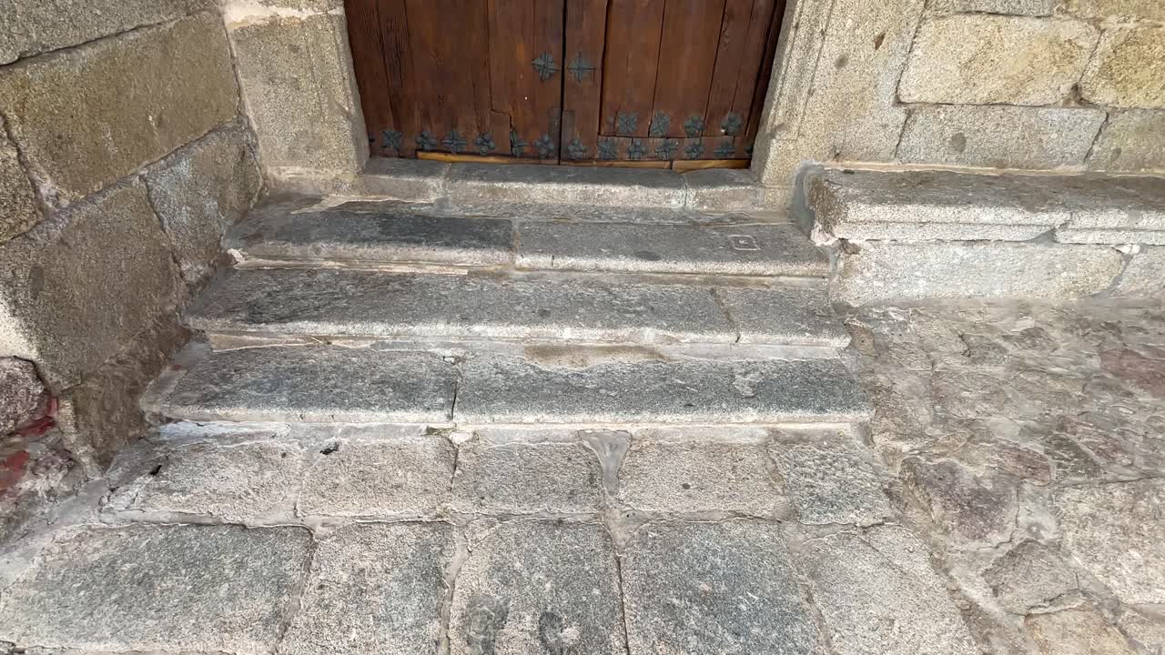 We see an old very worn white granite stone floor, a few steps away from it appears a spectacular large two-leaf wooden door, medieval style with its decorative iron studs, it is in slow motion