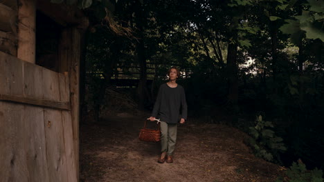 young person carrying a basket through a wooded area near a rustic building