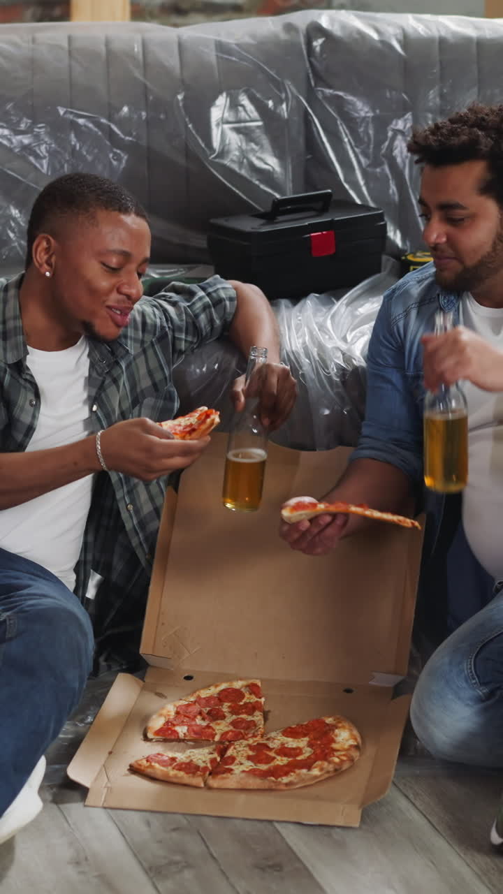African-American brothers with beer bottles eat fresh pizza sitting on floor by foiled sofa. Young men have rest after repairing works in living room