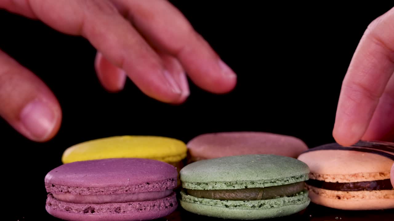 A hand carefully picks up a yellow macaron from a neat row of assorted colorful macarons on a glossy black surface under bright, even lighting