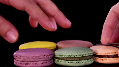a hand carefully picks up a yellow macaron from a neat row of assorted colorful macarons on a glossy black surface under bright, even lighting