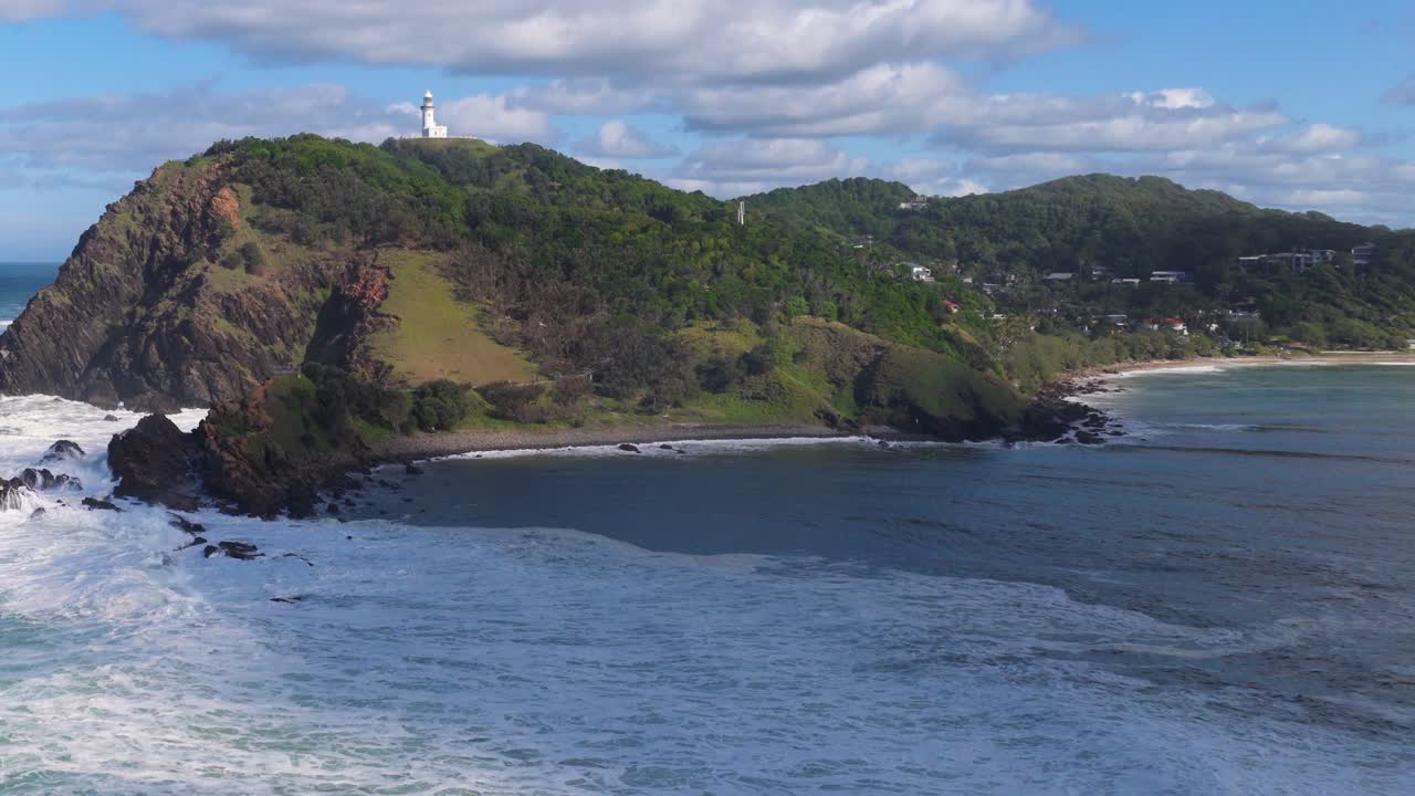 Drone footage captures Byron Bay's lighthouse atop a lush cliff, surrounded by ocean waves under a bright sky