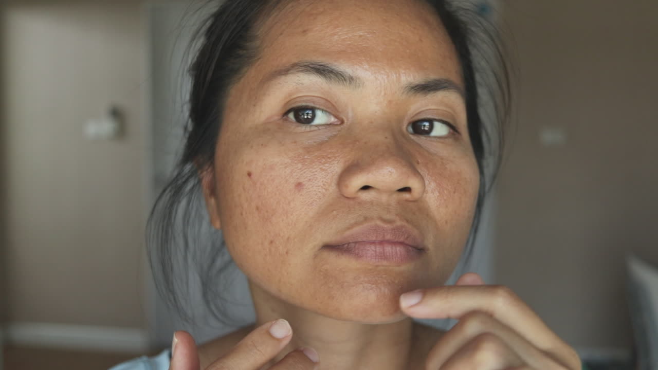 Primo piano di una donna che esamina acne e imperfezioni del viso usando entrambe le mani, girando la testa per controllare entrambe le guance. Ispezione mirata, luce naturale e texture realistica. Concetto di cura della pelle e dermatologia.