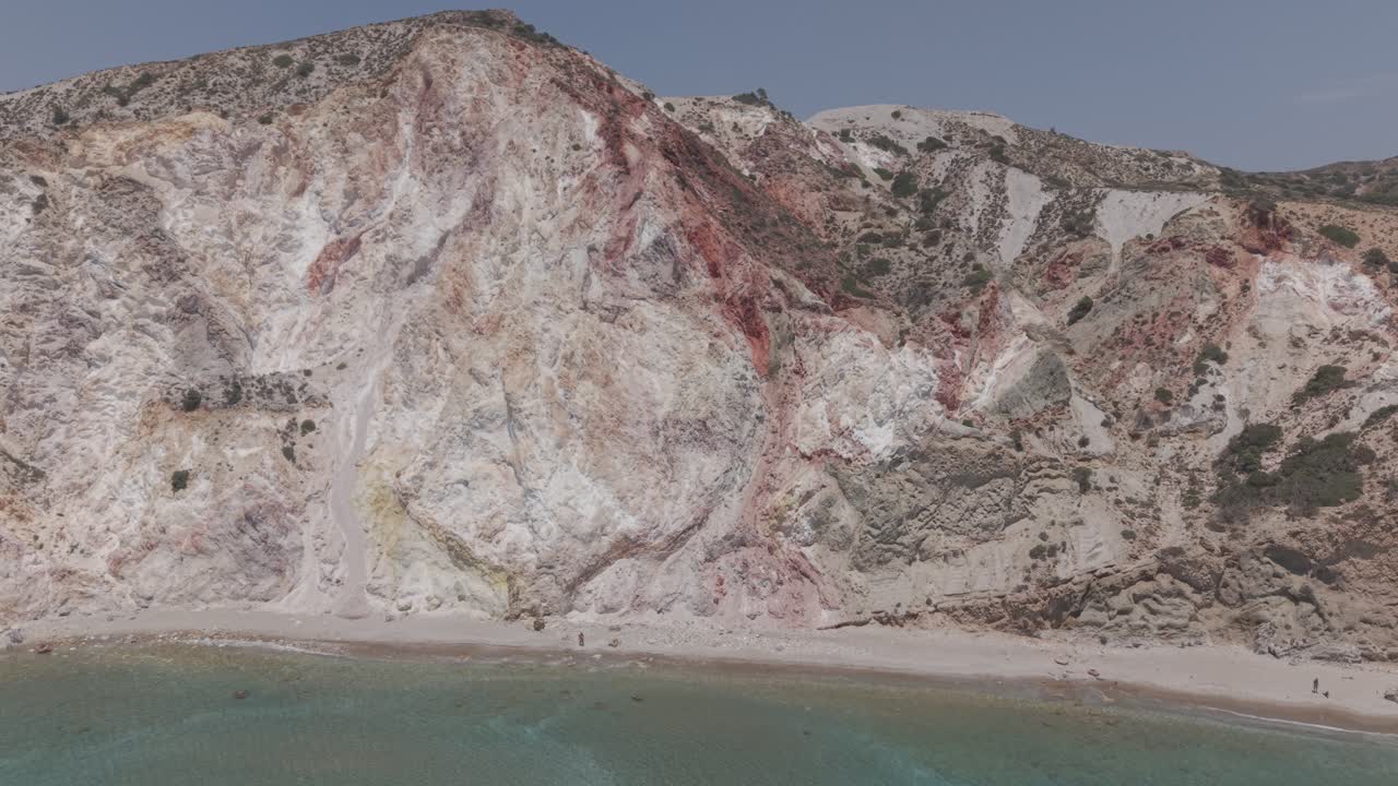 Vibrant cliffs in white, pink, yellow, and red rise above calm turquoise waters of the Aegean Sea under a bright blue sky on Milos Island, Greece. HD stock video