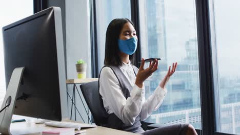 mujer asiática con máscara facial hablando en un teléfono inteligente mientras está sentada en su escritorio en una oficina moderna