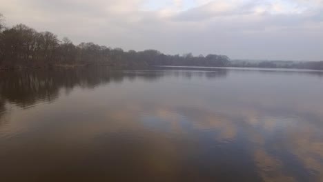 Flying-low-over-misty-mirrored-lake-with-woodland-to-left-of-shot