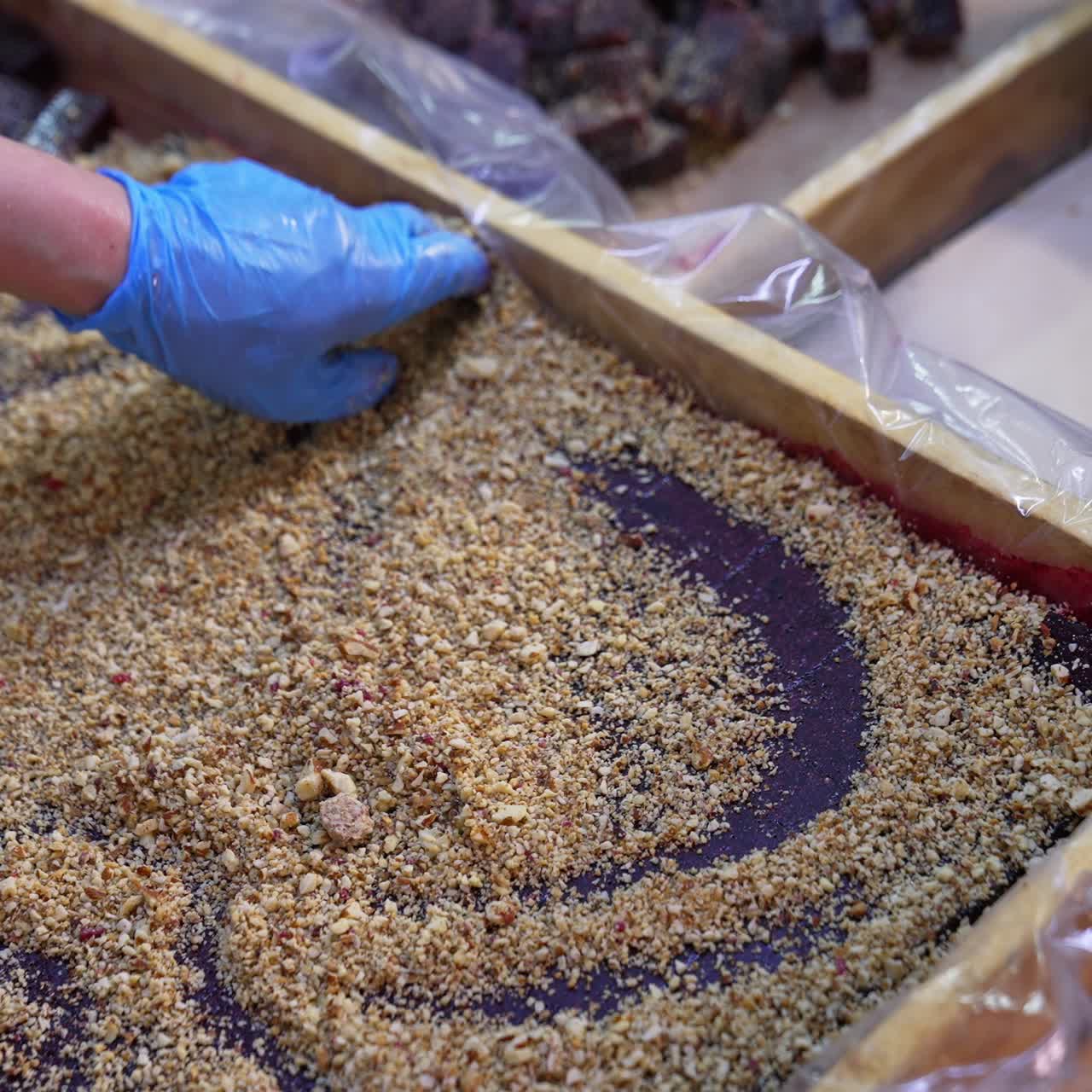 Purple dark layer of marmalade cut into pieces in a wooden box. Worker spreads nuts over the sweet candied fruit jelly. Close up