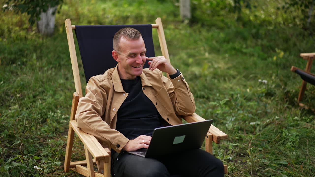 Caucasian man in brown shirt sitting with laptop on his laps in the garden. Relaxed freelance worker talks to someone laughing cheerfully.
