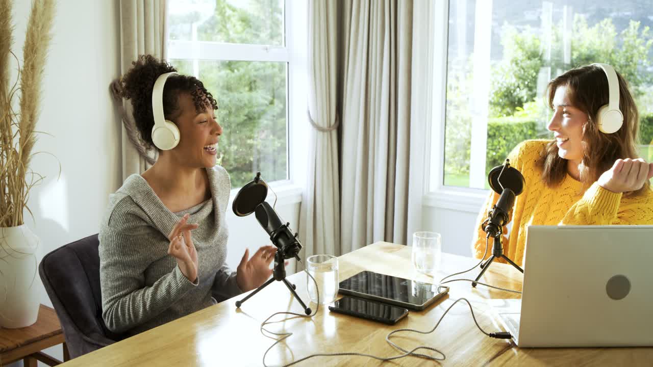 Compañeras de trabajo diversas levantando y ajustando auriculares, grabando un podcast con micrófonos en una mesa de estudio.