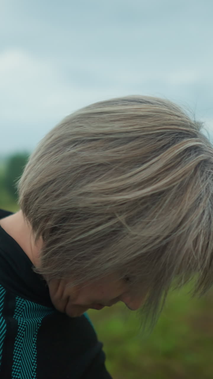 검은색과 초록색 의상을 입은 여성의 측면 뷰, 눈이 닫혀, 조용하고 조용한 야외 환경에서 그녀의 머리를 오른쪽에서 왼쪽으로 부드럽게 기울이고, 그녀는 긴장을 풀기 위해 주의 깊은 목 스트레칭을 연습합니다.